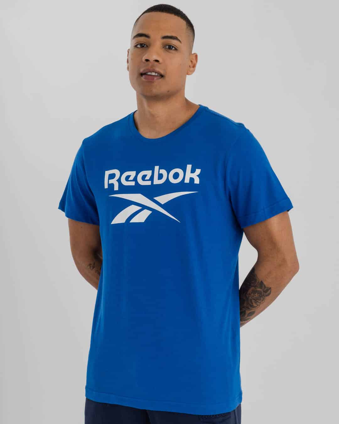 Full length shot of man wearing Blue Reebok t-shirt with Reebok Logo on chest in white & navy blue shorts.